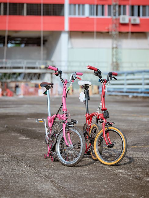 Two children's bicycles are positioned upright on a paved outdoor surface, with the pink bicycle on the left and the red bicycle on the right, both equipped with training wheels, handlebars, and seats