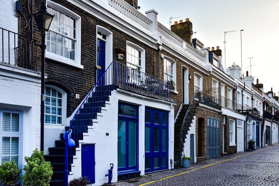Exterior view of a row of terraced houses on Brompton Road, SW3, showcasing traditional brick facades with white window frames and doors. Each house features small stairs leading up to the front doors, with some staircases painted in vibrant blue, including handrails and door frames, indicative of house entrances prepared for a home relocation or furniture transport. Outside, on the cobblestone pavement, there are potted plants and decorative elements. One house has a large glass panel door on the ground floor, with a smaller window above, and another door is accessed via the external staircase. The scene suggests the process of house removals, with visible loading areas suitable for moving boxes, furniture, and appliances. The lighting indicates daylight, providing clear visibility of all structural details. The setting is typical of the London residential streetscape where professional removals companies like Removal Companies Brompton facilitate packing and moving services, including loading and transport of household goods.