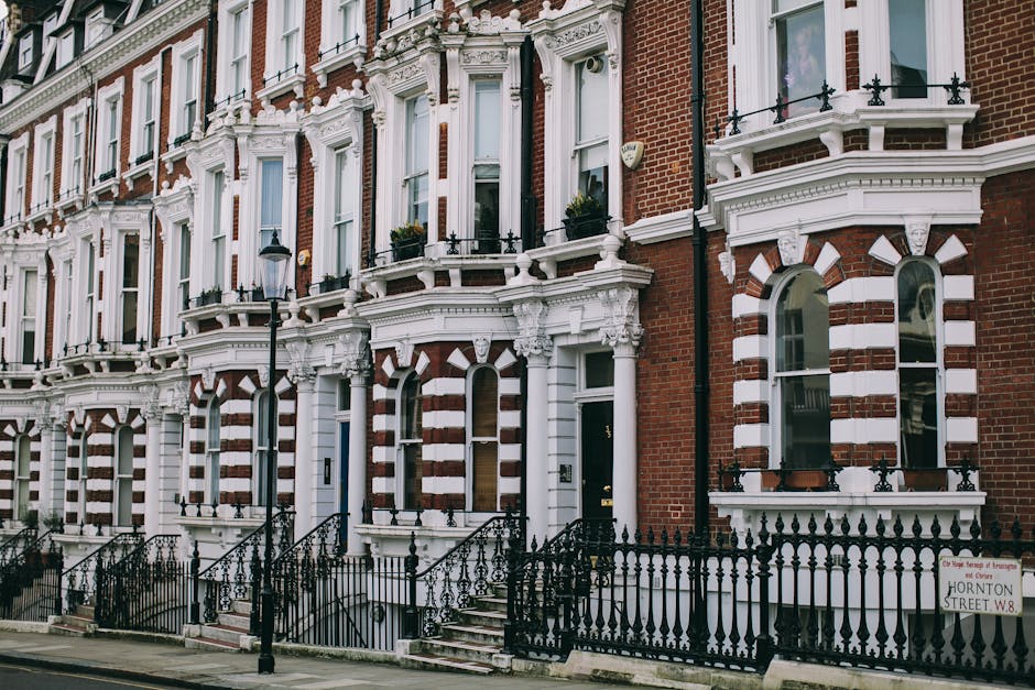 A row of Victorian-era terraced houses on Brompton Road, SW3, featuring red brick facades contrasted with white decorative architectural details around arched windows and bay windows. The ground floors have black wrought iron railings enclosing small front gardens and steps leading to black front doors, some of which have brass handles and modern doorbells. Black street lamps line the pavement in front, with a street sign indicating 'Hornton Street W8' visible near the bottom right. The image is taken during daylight hours, showcasing the exterior environment where house removals or furniture transport may occur, with no visible vehicles or moving equipment present in this specific scene.