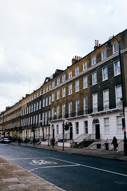 Exterior view of a row of terraced houses on Brompton Road, SW3, showcasing traditional brick facades with white window frames and doors. Each house features small stairs leading up to the front doors, with some staircases painted in vibrant blue, including handrails and door frames, indicative of house entrances prepared for a home relocation or furniture transport. Outside, on the cobblestone pavement, there are potted plants and decorative elements. One house has a large glass panel door on the ground floor, with a smaller window above, and another door is accessed via the external staircase. The scene suggests the process of house removals, with visible loading areas suitable for moving boxes, furniture, and appliances. The lighting indicates daylight, providing clear visibility of all structural details. The setting is typical of the London residential streetscape where professional removals companies like Removal Companies Brompton facilitate packing and moving services, including loading and transport of household goods.