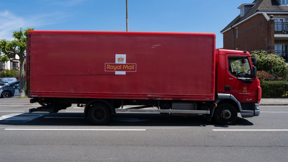 A red Royal Mail delivery truck is parked on the side of a residential street during daytime. The truck has a large, enclosed cargo area with the Royal Mail logo prominently displayed on the side, featuring a crown atop a white background and the words 'Royal Mail' in yellow. The vehicle is positioned parallel to the pavement, with the driver’s cab facing to the right. Behind the truck, there are multi-storey brick residential buildings with balconies, and trees with green foliage lining the street. The scene is well-lit with clear blue sky overhead. This image is relevant to house removals and furniture transport, illustrating the use of delivery vehicles in relocation logistics, as handled by companies such as Removal Companies Brompton.
