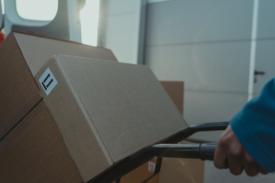 Close-up of a person wearing a blue sleeve pushing a black hand truck loaded with several cardboard boxes, some of which are sealed with packing tape, inside a building with natural light coming through a window. The boxes are arranged in a stack, with larger boxes at the bottom and smaller ones on top, indicating a home relocation process. In the background, there is a white door and a partially visible wall, suggesting the interior of a property preparing for furniture and household items transport. This scene reflects the packing and loading phase of residential removals, supported by Removal Companies Brompton for house moves and relocation logistics.
